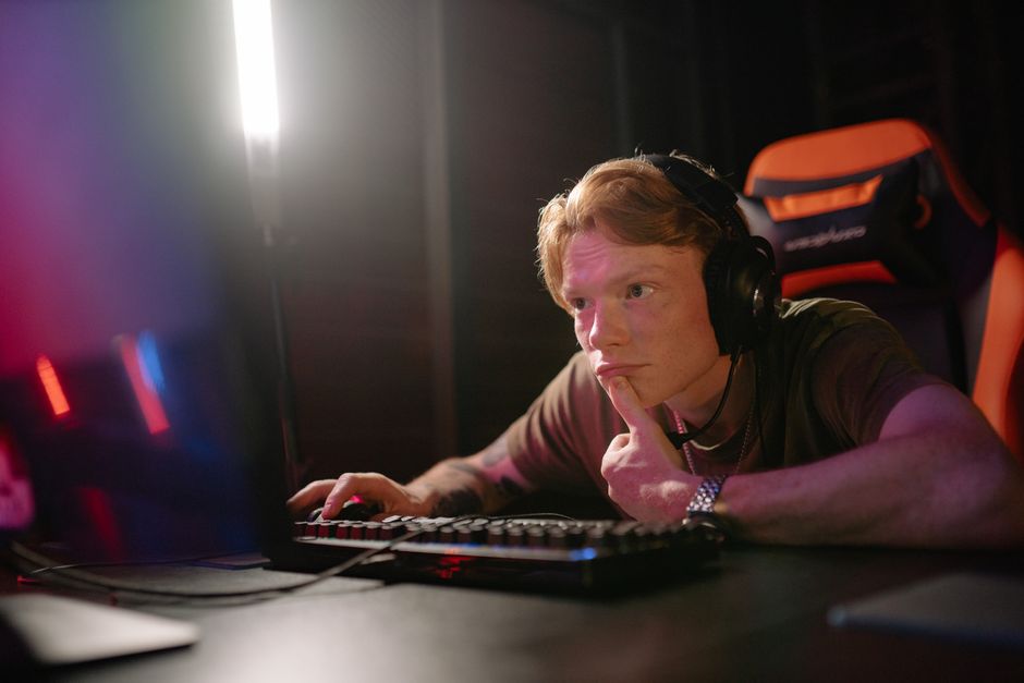 A young man intensely gaming on a computer, surrounded by tech gear in a dimly lit room.