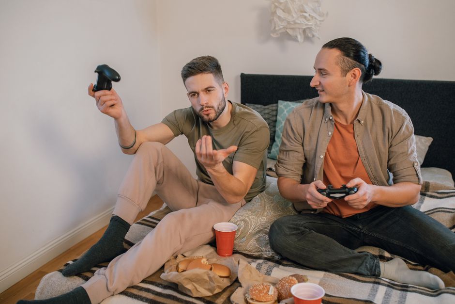 Two men sitting on a bed playing video games with controllers, enjoying leisure time.