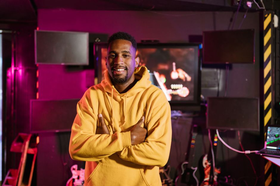 Smiling gamer in a yellow hoodie stands confidently in an e-sports studio with vibrant lighting.